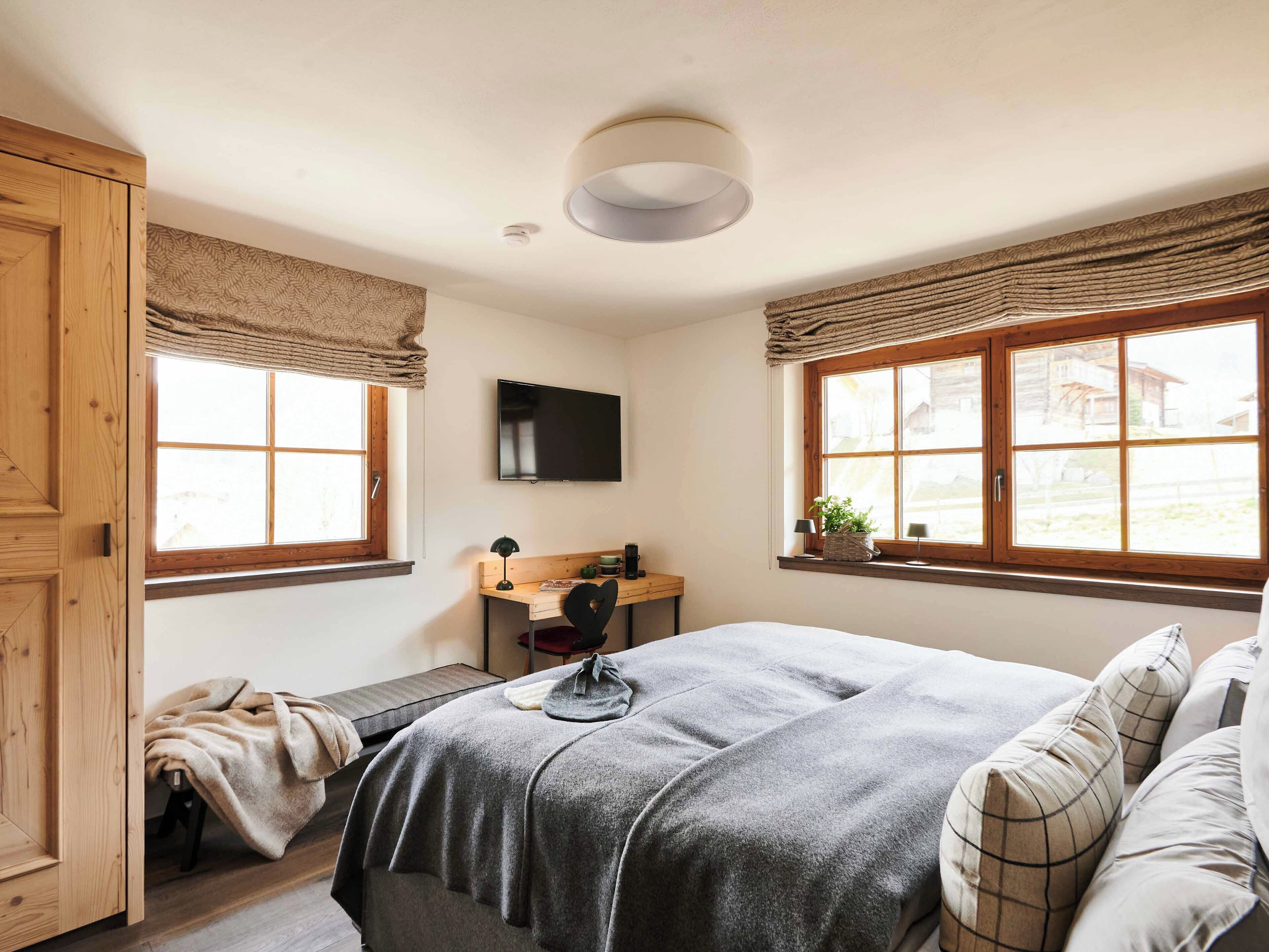 A bedroom with a bed, a desk, a chair, a television, and windows with curtains.