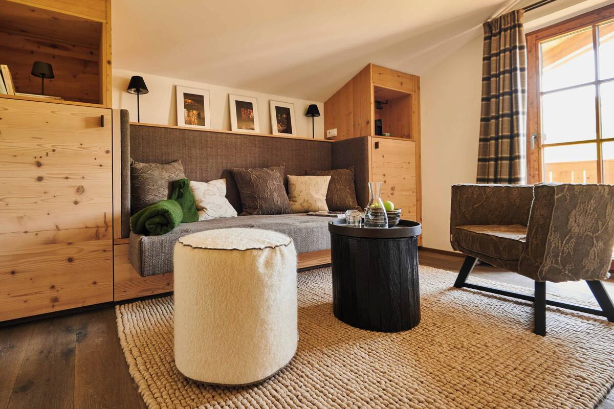 Una sala de estar acogedora con sofá, reposapiés y sillón, armarios de madera y un amplio ventanal con cortinas.