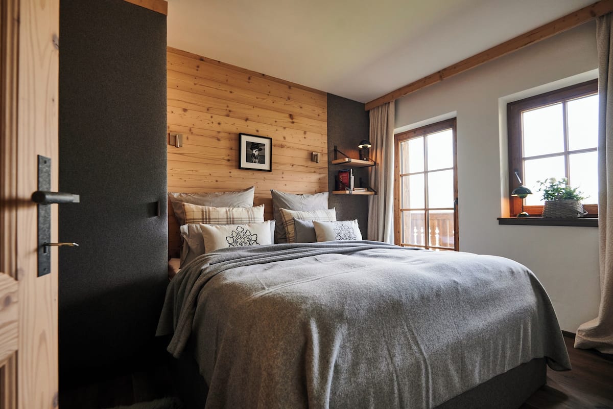 Un dormitorio con una cama, almohadas, cabecera de madera, una ventana con cortinas y una estantería con libros y una lámpara.