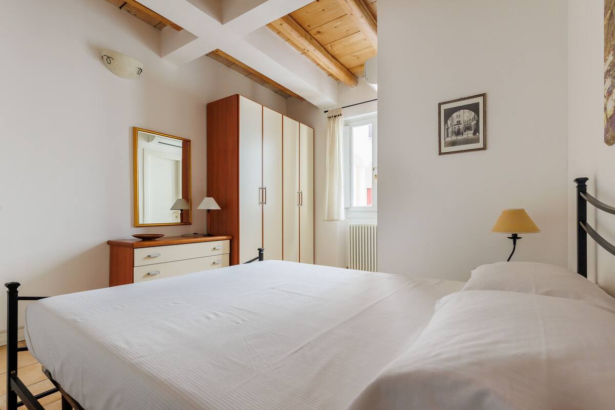 A bedroom with a bed, a dresser, a mirror, a window with curtains, and a wall-mounted lamp.