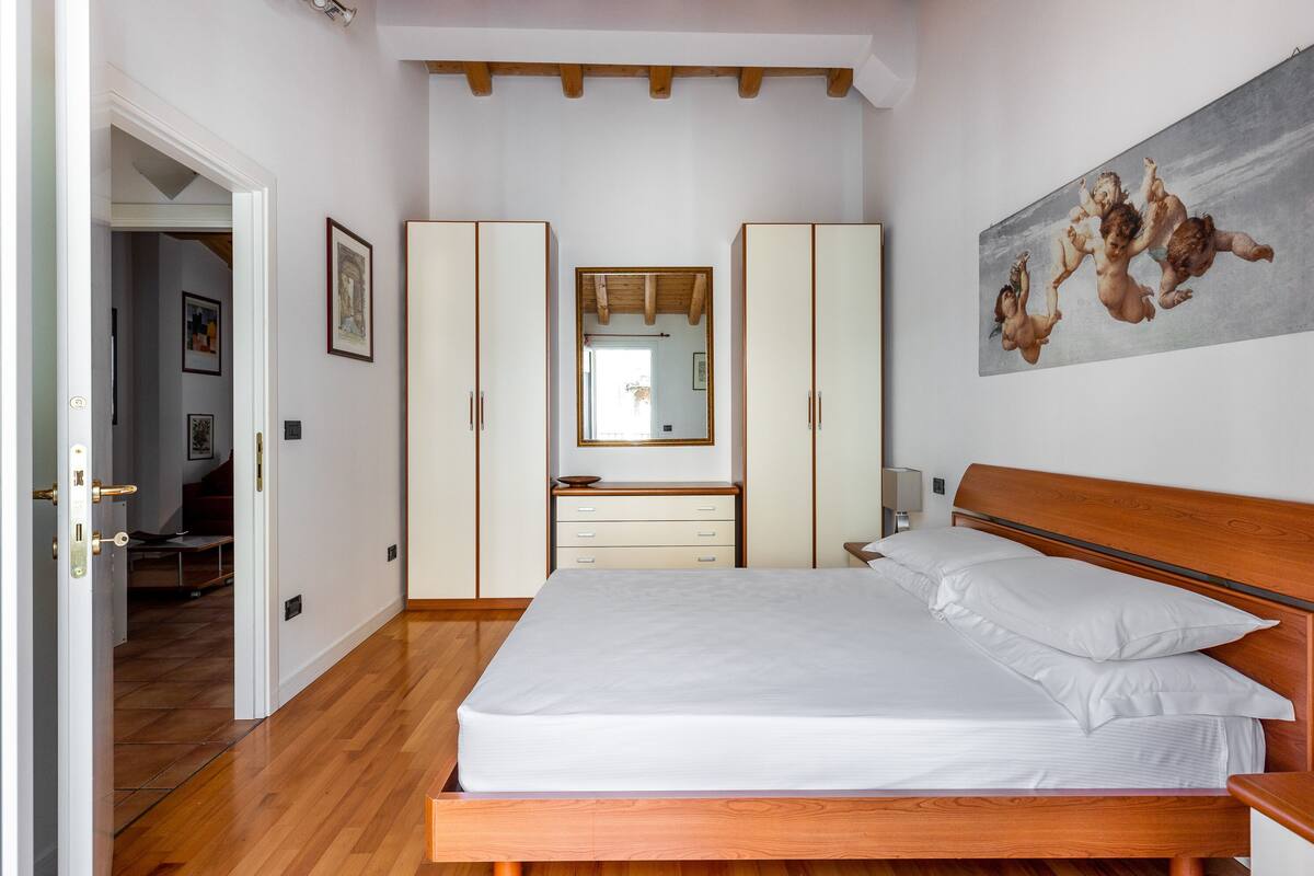 A bedroom with a wooden bed, white bedding, a wooden headboard, a large mirror, and a painting on the wall.