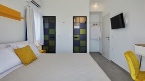 A hotel room with a bed, a yellow chair, a TV, and a bathroom with a towel rack. - Thadeos Villa (Santorini)