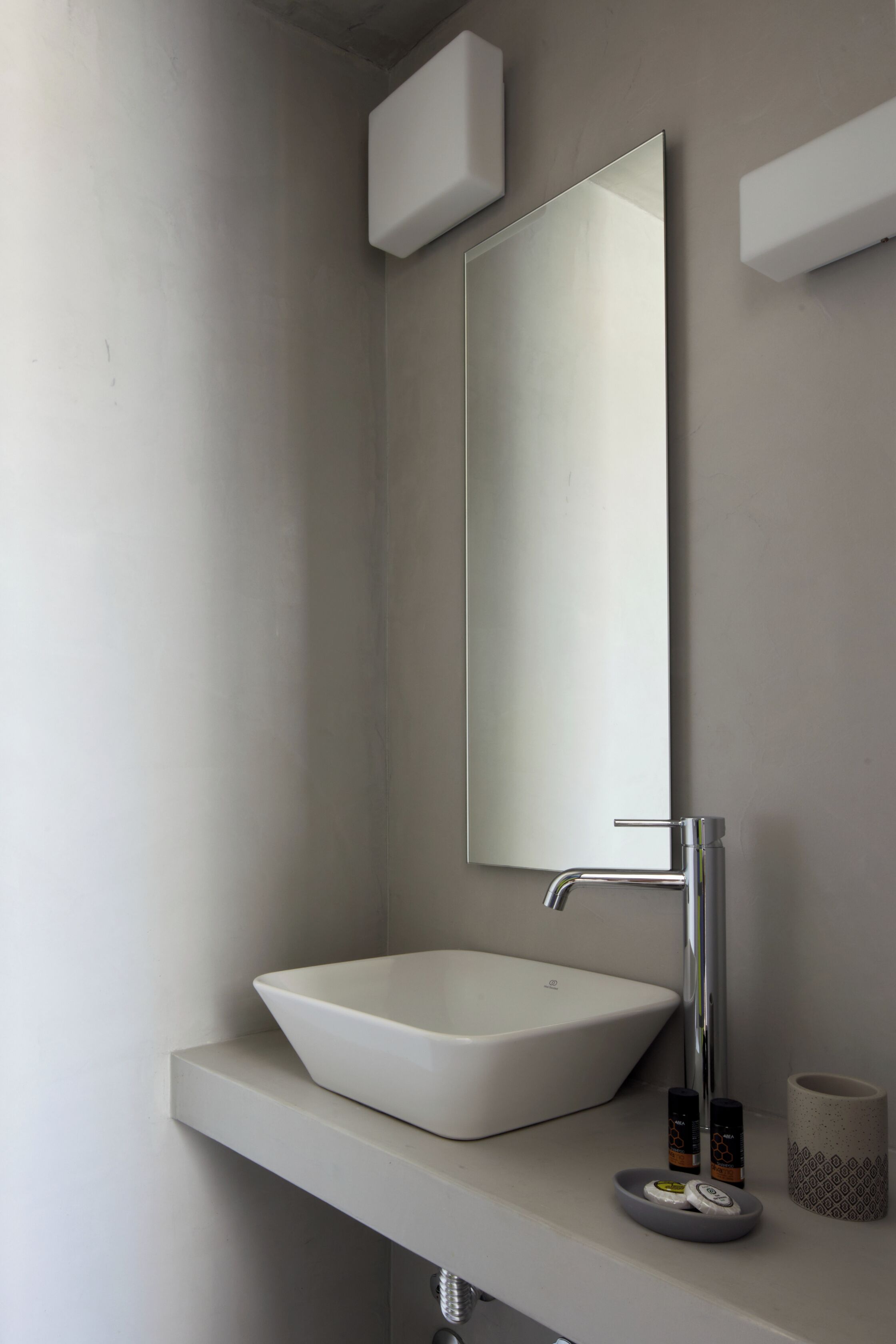 A modern bathroom with a white sink, a rectangular mirror, and a small shelf with toiletries.