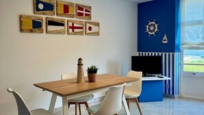 A dining area with a wooden table and white chairs, a wall with nautical-themed artwork, and a blue accent wall with a TV and a small sailboat decoration. - Preciosa Vivienda Para Vacaciones a 50m de la Playa de Altar (Barreiros)