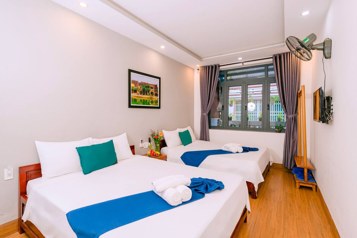 A hotel room with two beds, a large window with curtains, a standing fan, and a picture on the wall.