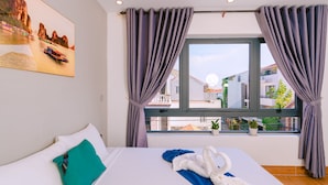 A hotel room with a bed, a window with curtains, a painting on the wall, and a view of buildings outside. - Vinh Khang Homestay (Da Nang)