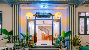 A hotel entrance with a sign reading "VINH KHANG," potted plants, a staircase, and a reception desk. - Vinh Khang Homestay (Da Nang)
