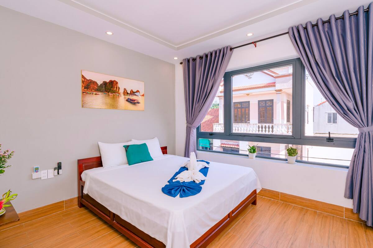 A hotel room with a neatly made bed, a window with curtains, a painting on the wall, and a view of the outside buildings.