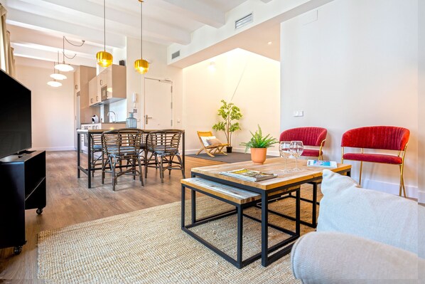 A modern living room with a wooden coffee table, a sofa, and red upholstered chairs. There is a dining area with a wooden table and chairs, and a kitchen in the background.