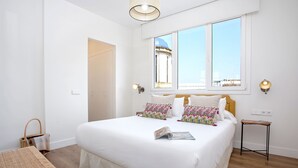 A spacious bedroom with a large bed, a wooden table, and a view of a building through the window. - Genteel Home Almirante Lobo II (Sevilla)