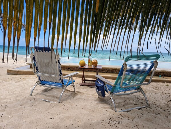 Two beach chairs with a table between them, a bottle, and a coconut on the sand near the ocean. - A la orilla del mar ok2 (Chuburná)