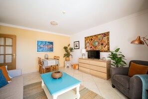 Uma sala de estar com sofá, mesa de centro azul, televisão, área de jantar com mesa e cadeiras, e cozinha com armário e uma planta.