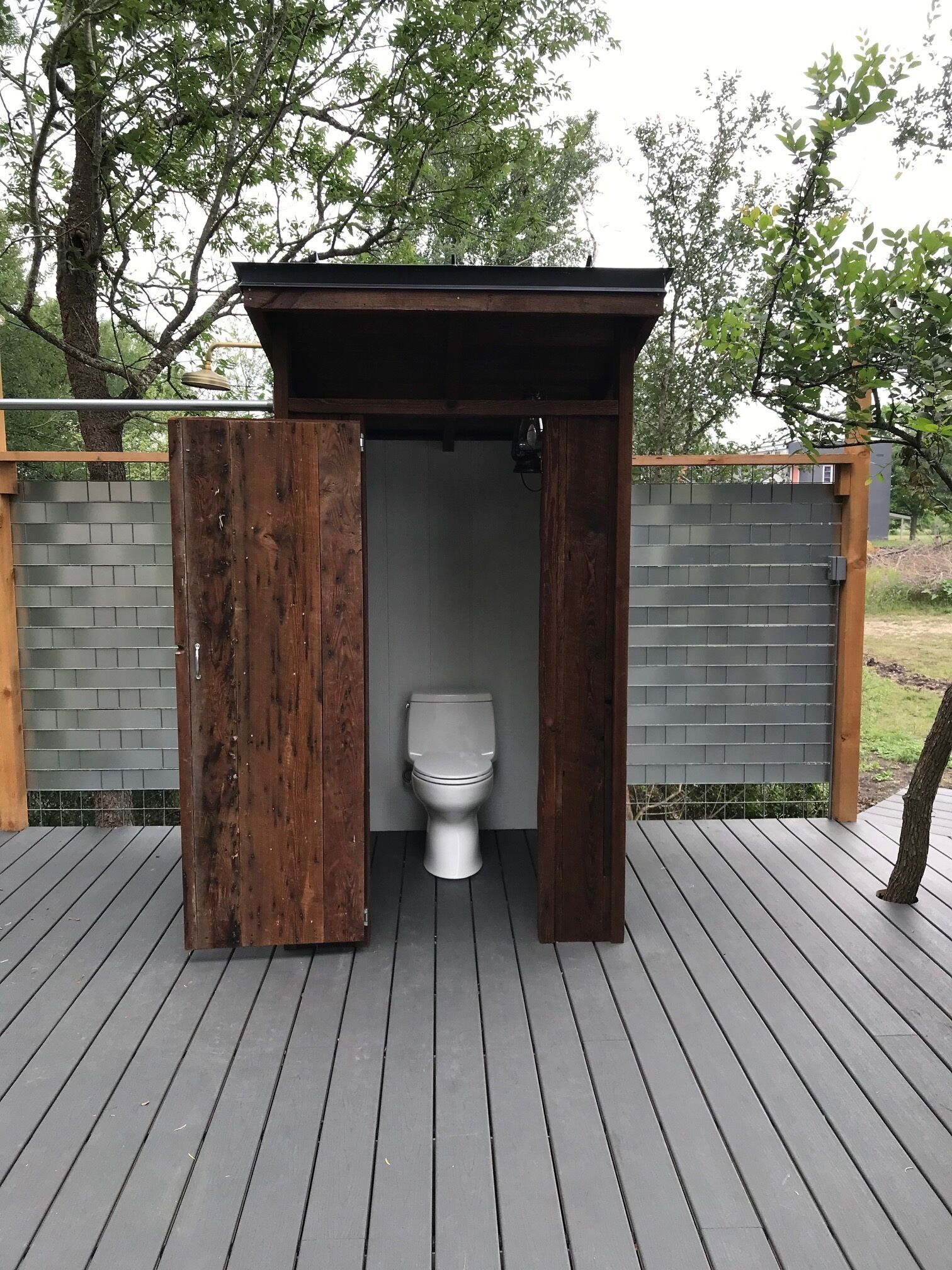 A wooden outdoor toilet with a metal roof, surrounded by a wooden deck and a brick wall.