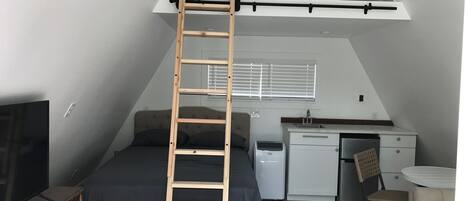 Ein kleines Zimmer mit Schlafkoje, einer Holzleiter, einer Küchenzeile, einem Fernseher und einem kleinen Esstisch.