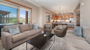 A spacious living room with a large sofa, two armchairs, and a coffee table. A kitchen with wooden cabinets and a dining area is visible in the background.