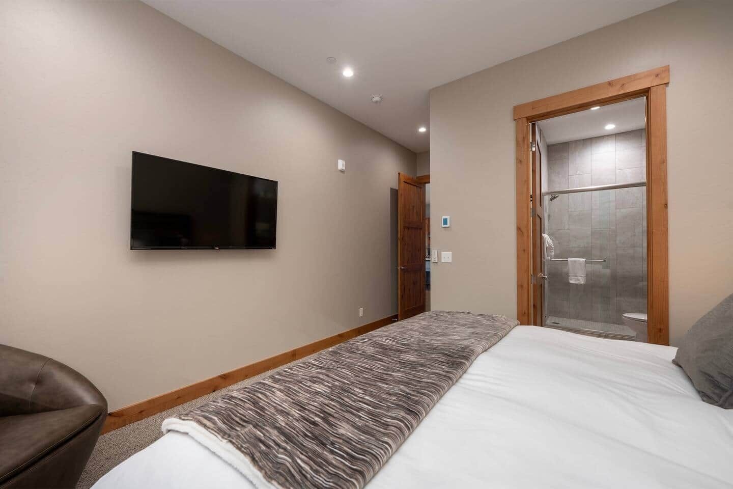 A bedroom with a bed, a TV mounted on the wall, a shower area, and a door leading to another room.
