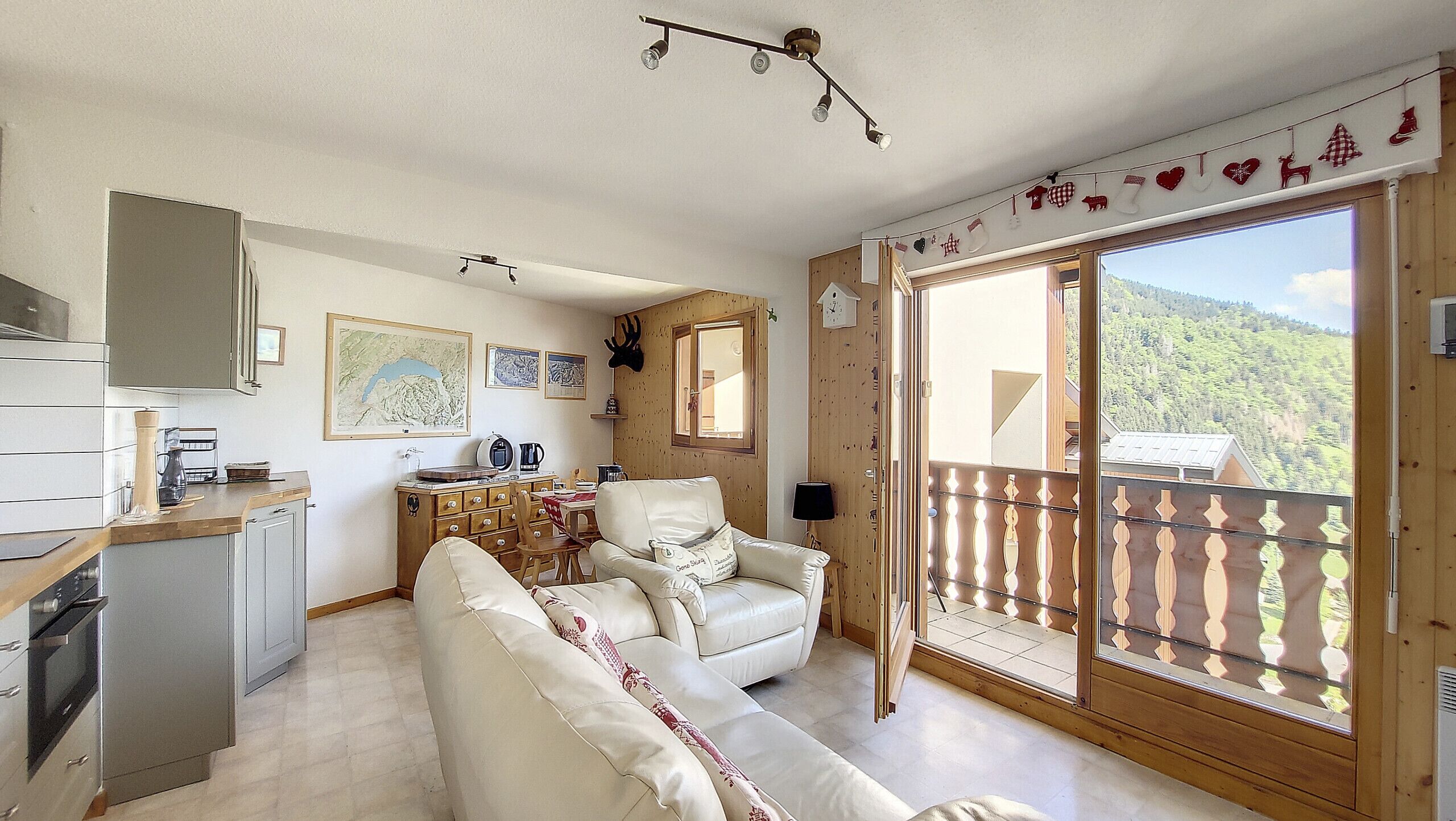 A cozy room with a sofa, a dining table, and a kitchen area. There is a balcony with a wooden railing and a view of the mountains.