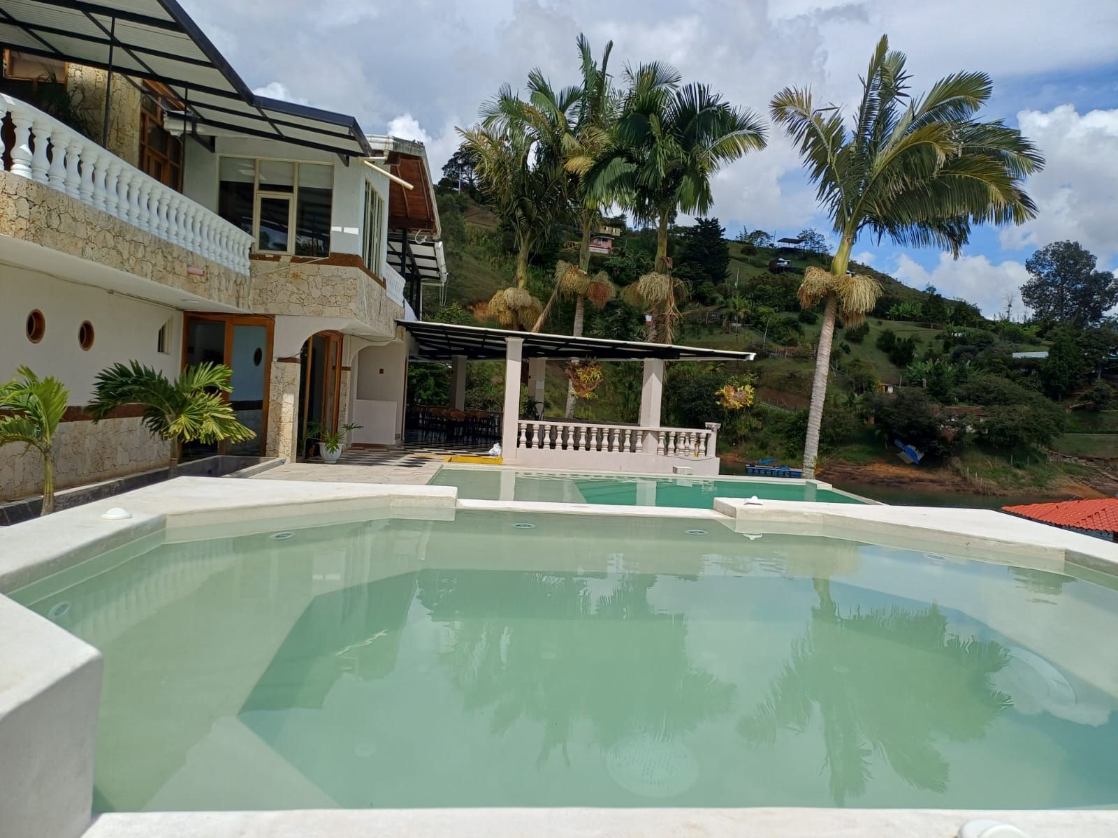 Una piscina con zona cubierta, una casa con balcón y palmeras al fondo.