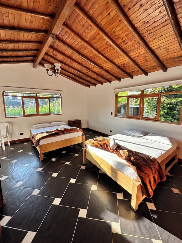 A room with two wooden beds, a white chair, and large windows with a view of greenery. - Casa Linda Guatapé (Guatapé)