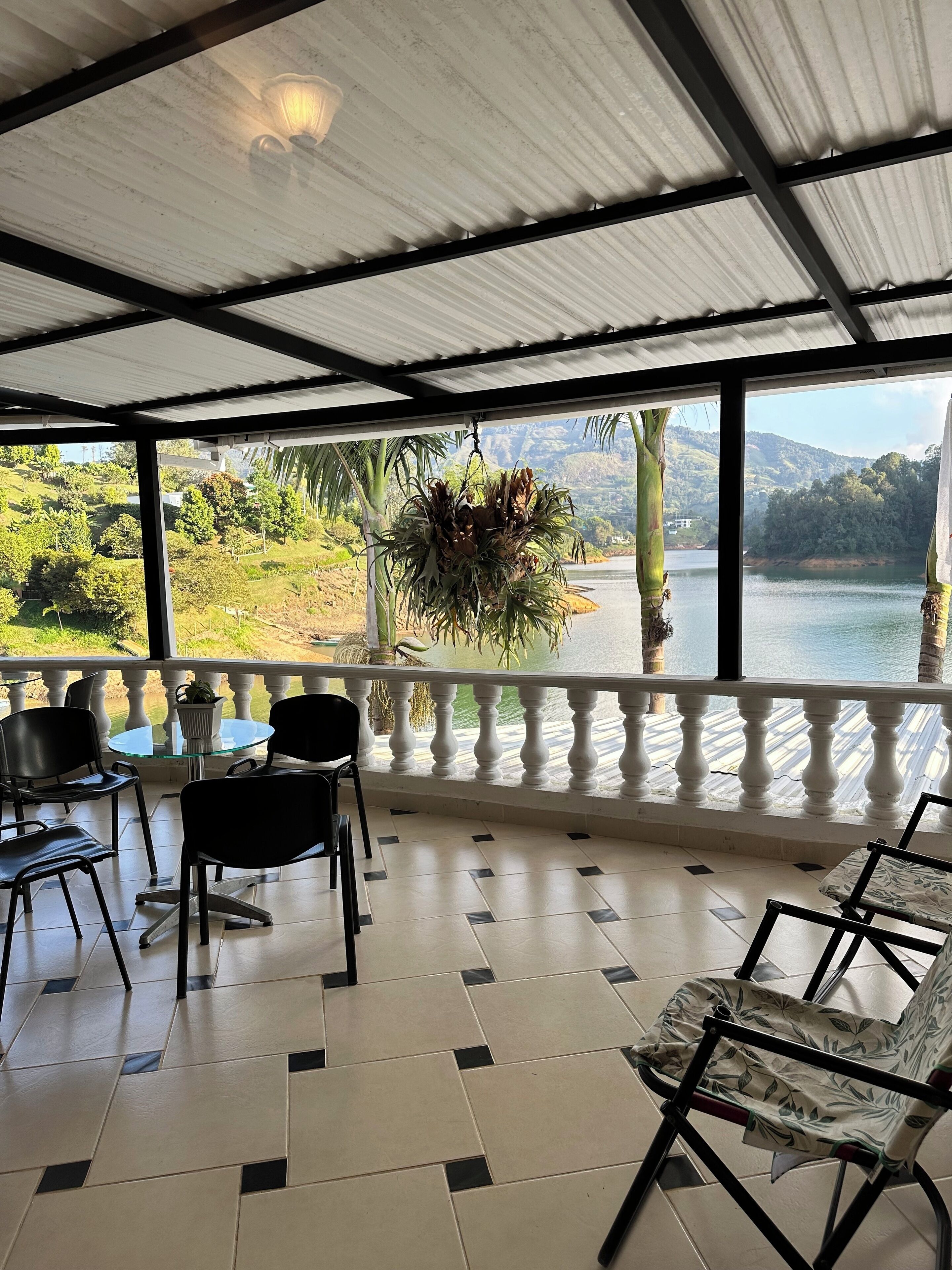 A covered terrace with a view of a lake and hills, furnished with chairs and a table.