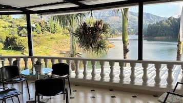 A covered terrace with a view of a lake and hills, furnished with chairs and a table.