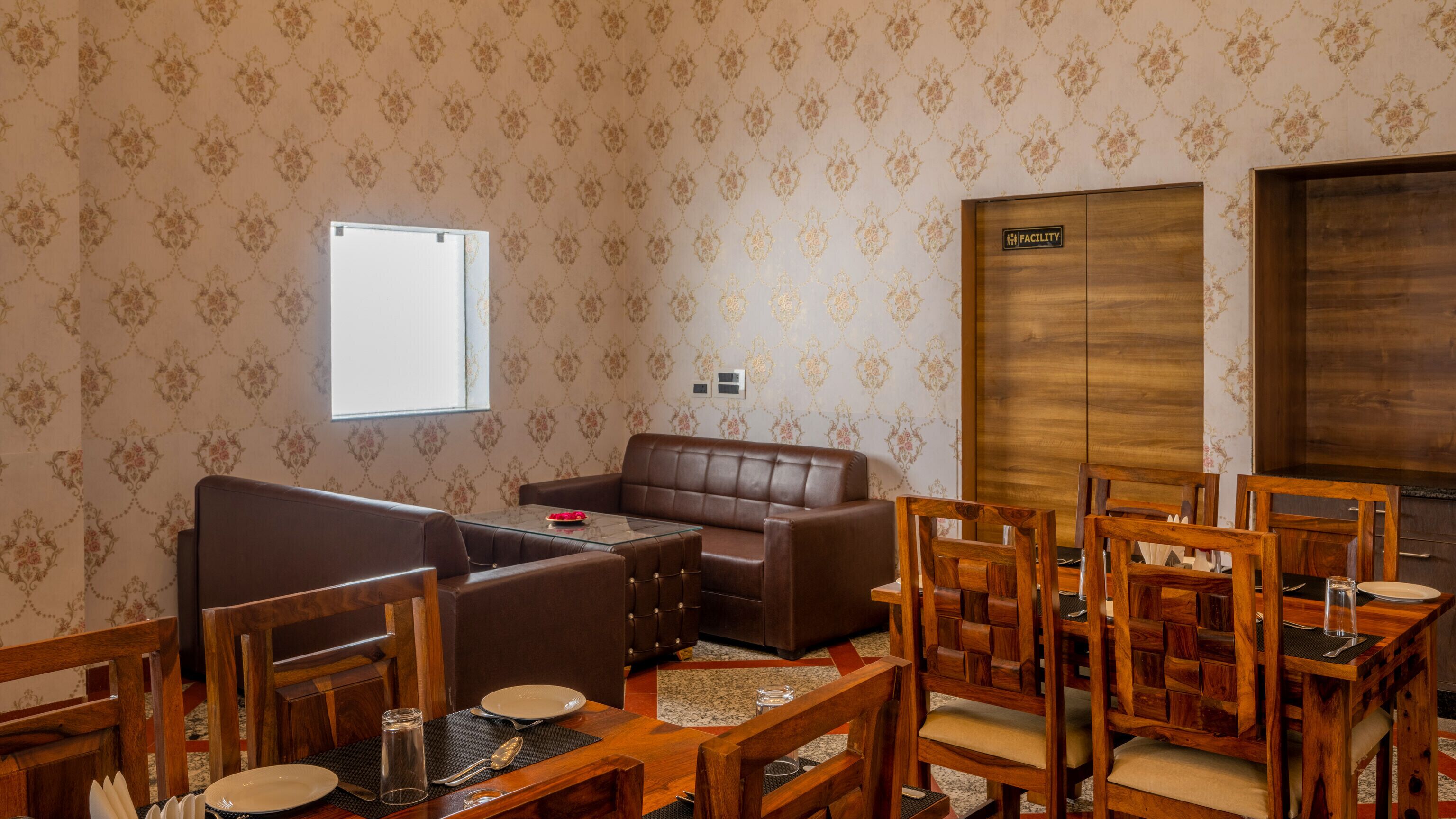 A hotel room with a brown leather sofa, wooden dining table with chairs, a small glass table, and a large wooden wardrobe.