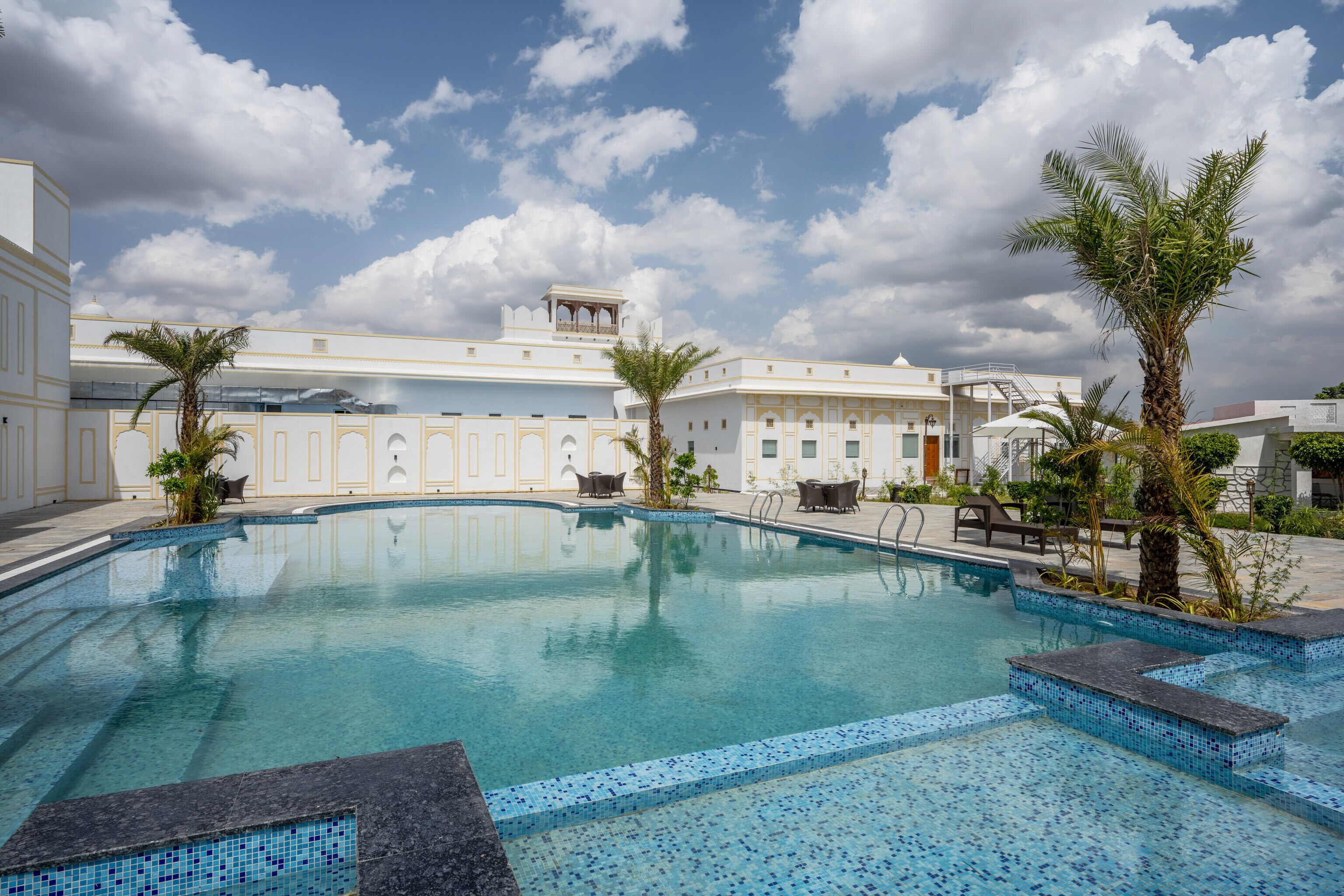 A swimming pool with tiled edges, surrounded by palm trees and a building with a white facade and arched windows.