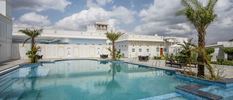 A swimming pool with tiled edges, surrounded by palm trees and a building with a white facade and arched windows.