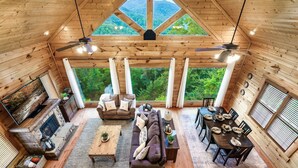 A spacious cabin with a stone fireplace, a large sofa, a dining table set for six, and large windows offering a view of the forest. - Mega Views! 6 Miles to GSMNP! Hot Tub, Pool Table, Arcade, Foosball, 2Kg, 2QN (Sevierville)