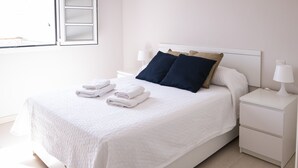 Un lit bien fait, avec du linge de lit blanc et deux oreillers, une table de chevet blanche avec une lampe, et une fenêtre avec des stores.