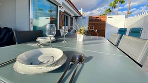 Een terras met eettafel voor twee personen, een potplant en uitzicht op palmbomen en een gebouw.