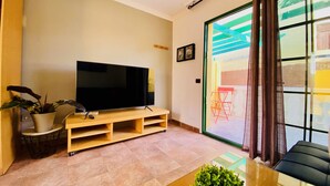 A living room with a flat-screen TV on a wooden stand, a potted plant, and a glass coffee table.