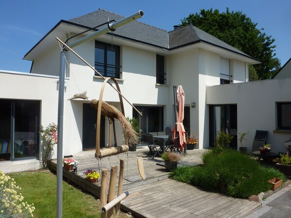 A modern two-story house with a patio area featuring a table and chairs, a hanging chair, and potted plants.