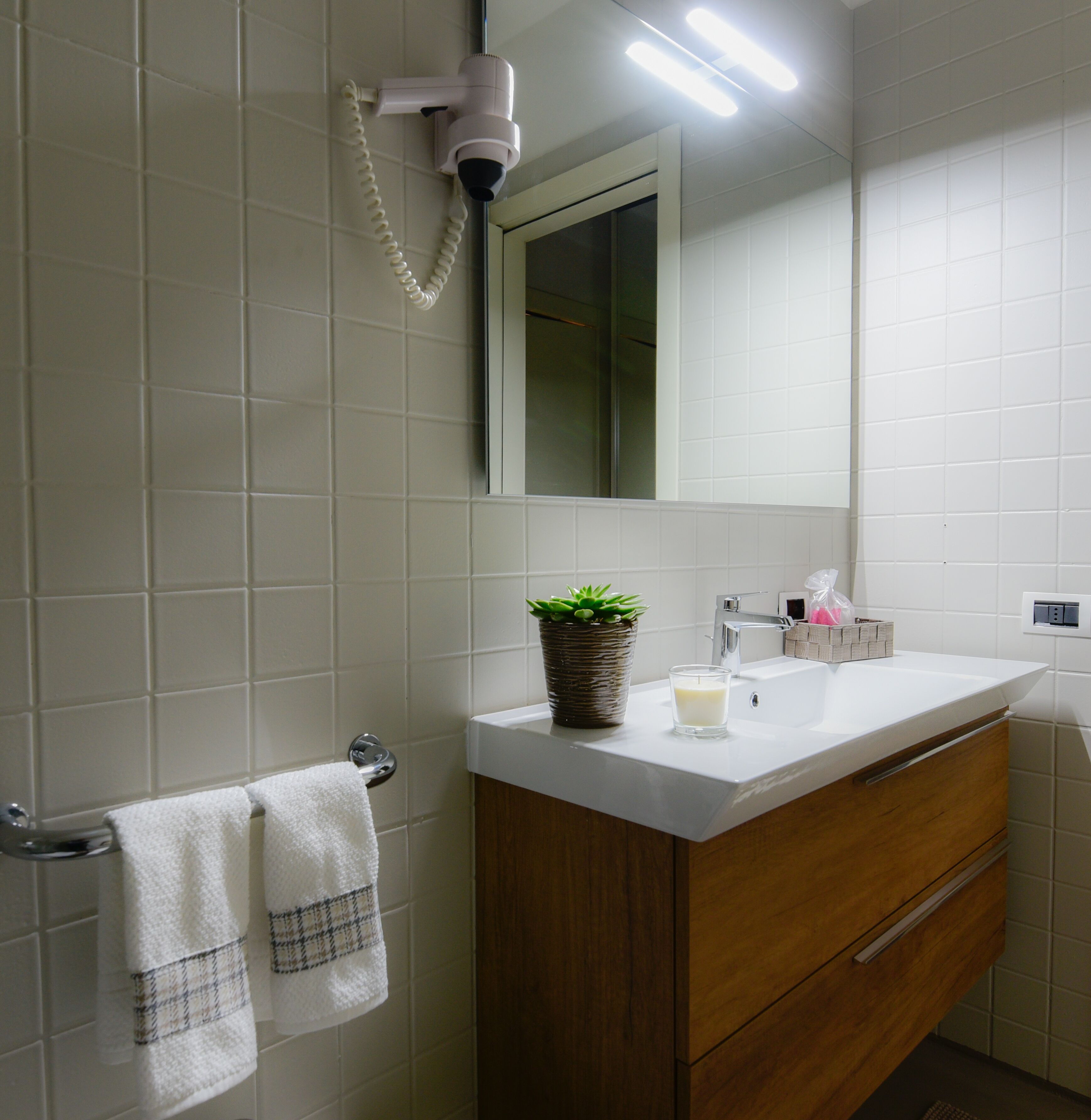 Une salle de bain avec des carreaux blancs, un meuble-lavabo en bois, un miroir, un sèche-cheveux, un lavabo, une bougie en verre et une plante en pot.