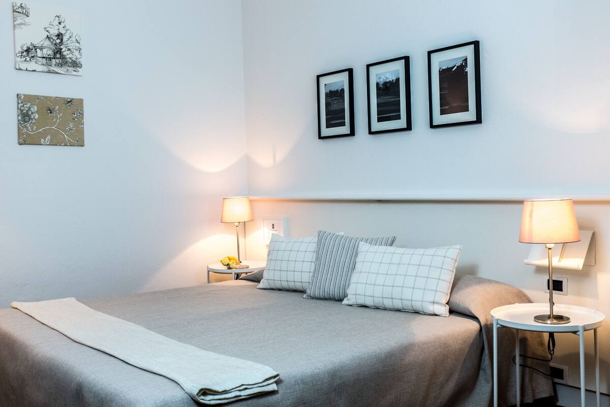Ein ordentlich gemachtes Bett mit grauer Tagesdecke und Kissen, zwei Nachttische mit Lampen und drei gerahmte Bilder an der Wand.