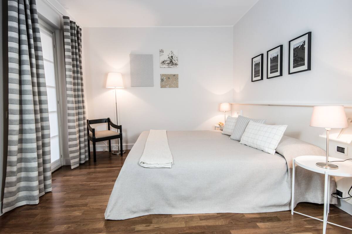 Ein ordentlich eingerichtetes Hotelzimmer mit einem großen Bett, zwei Nachttischen mit Lampen, einem Sessel und Blick ins Freie durch Vorhänge.