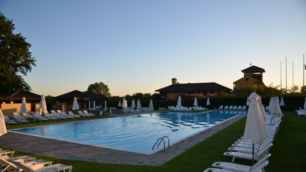 A swimming pool with lounge chairs and umbrellas, surrounded by well-maintained grass and a building with a tower. - Golf Hotel Castelconturbia (Agrate Conturbia)