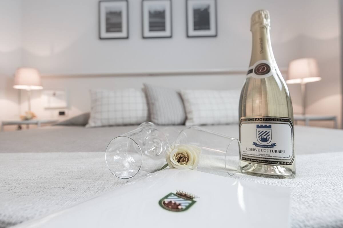 Eine Champagnerflasche und zwei Gläser auf einem Bett in einem Hotelzimmer, mit einem ordentlich bezogenen Bett, Kissen und gerahmten Bildern an der Wand.