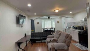 A compact living space with a sofa, a dining table, a TV mounted on the wall, and a kitchenette area.