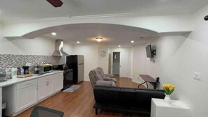 A modern kitchen with white cabinets, a microwave, and a stove. There is a dining area with a black sofa and a small table. A clock is visible on the wall.