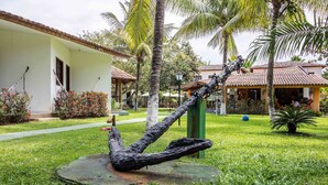 A sculpture of a figure in a relaxed pose on a circular platform in a grassy area with palm trees and buildings in the background. - Pousada Galeão Santa Anna (Maraú)