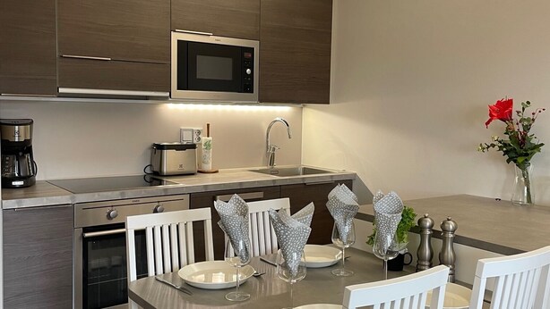 A modern kitchen with a built-in microwave, oven, and coffee maker. There is a dining table with white chairs and a vase with red flowers. - Spa Hotel Red Aparments (Kuopio)