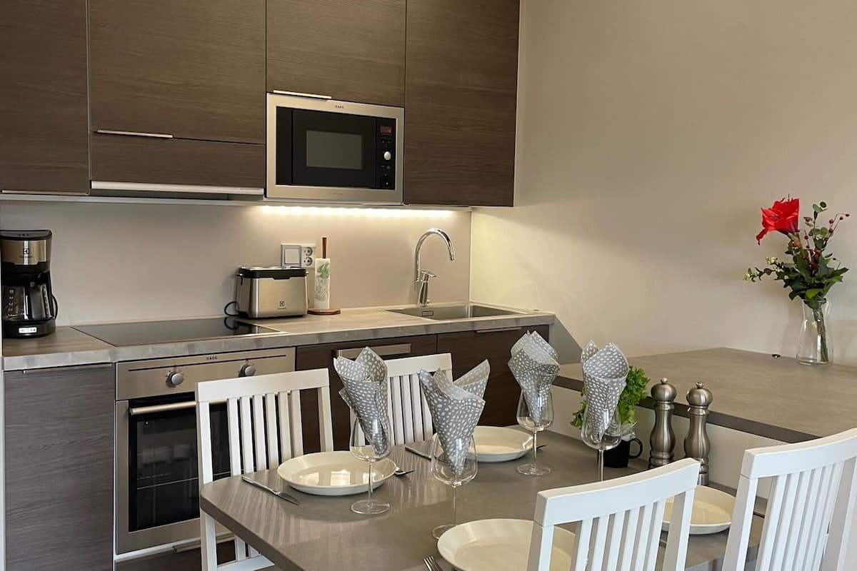 A modern kitchen with a built-in microwave, oven, and coffee maker. There is a dining table with white chairs and a vase with red flowers.
