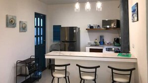 A compact kitchen with a central island, a refrigerator, a microwave, and a sink. There are three bar stools and a small dining table. The kitchen has a modern design with pendant lighting.