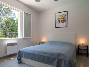 Une chambre avec un grand lit, deux tables de chevet avec des lampes, une fenêtre donnant sur des arbres et un tableau encadré au mur.