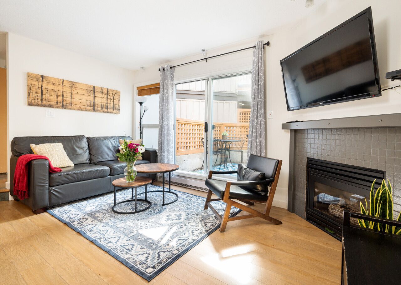 A modern living room with a black leather sofa, a round coffee table, a black armchair, a flat-screen TV mounted above a fireplace, and a large window with a view of a building outside.