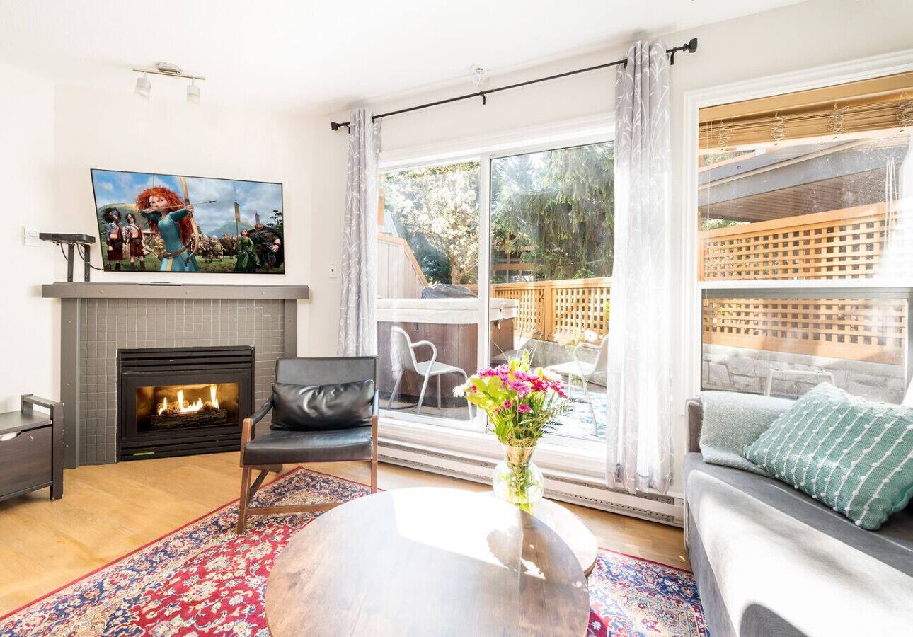 A modern living room with a fireplace, a large window with a view of the outdoors, a sofa, and a coffee table with a vase of flowers.