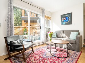 A living room with a sofa, a coffee table, a chair, a large window, and a decorative rug. - Private Hot Tub, Patio and Free Parking @ Glaciers (Whistler)