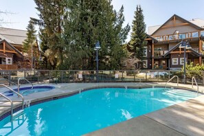 A swimming pool with a tiled edge, surrounded by a fence, with a building featuring a wooden facade and large windows in the background. - Private Hot Tub, Patio and Free Parking @ Glaciers (Whistler)