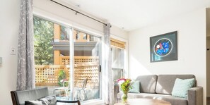 A living room with a sofa, a coffee table, and a chair. There is a large window with curtains, a balcony, and a framed picture on the wall.